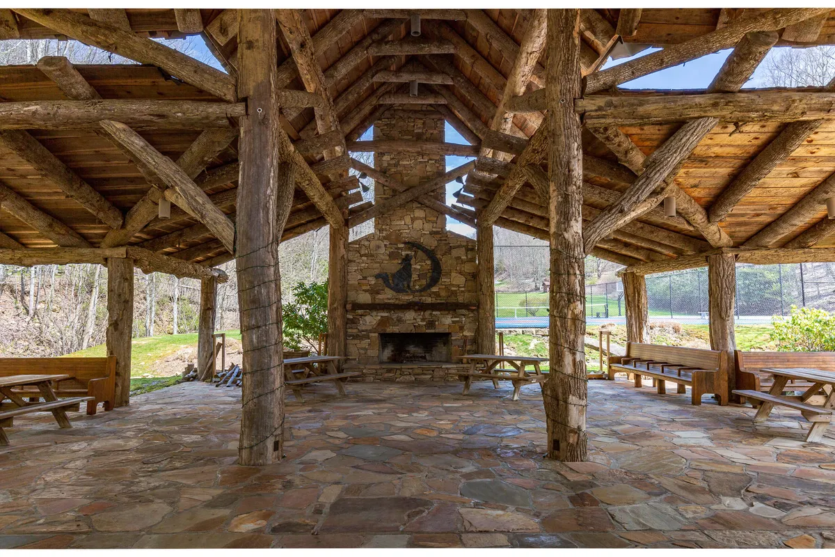 The scene highlights an outdoor communal area with a stone fireplace, wooden beams, picnic tables, and peaceful natural surroundings. The image evokes warmth, togetherness, and communal experiences, perfect for group celebrations and storytelling under the stars.