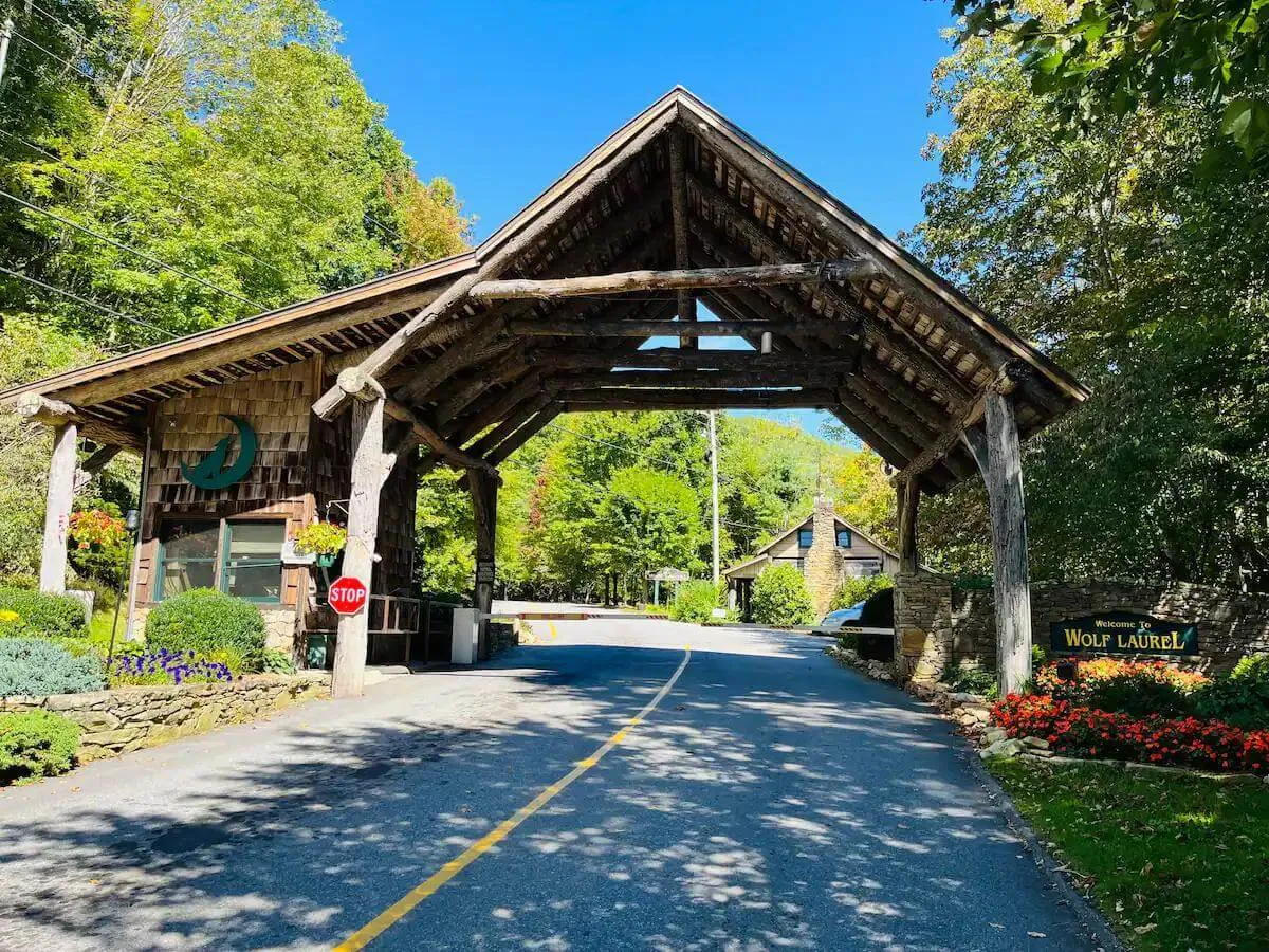 A grand wooden archway with a welcoming sign greets arriving guests, surrounded by flower beds and leading into a road flanked by greenery. The entrance feels both inviting and distinctly rustic, signaling arrival at a unique destination.