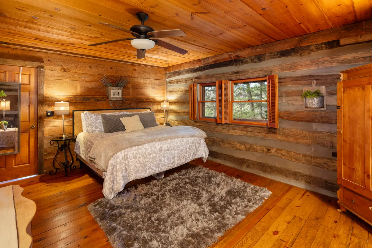 A cozy bedroom scene with a decorative bed, wooden wardrobe, windows with classic shutters framing woodland views, and a soft rug underfoot. Sunlight gently filters into the room, creating an inviting space ideal for rest or reading.
