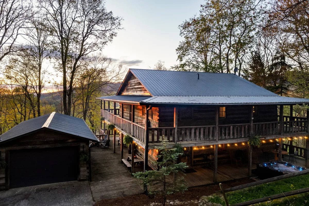 The image shows a beautifully crafted log cabin, warmly lit in the evening, with expansive wrap-around decks nestled among tall trees. The inviting porch hints at comfortable seating for gatherings while the surrounding nature exudes tranquility and seclusion.