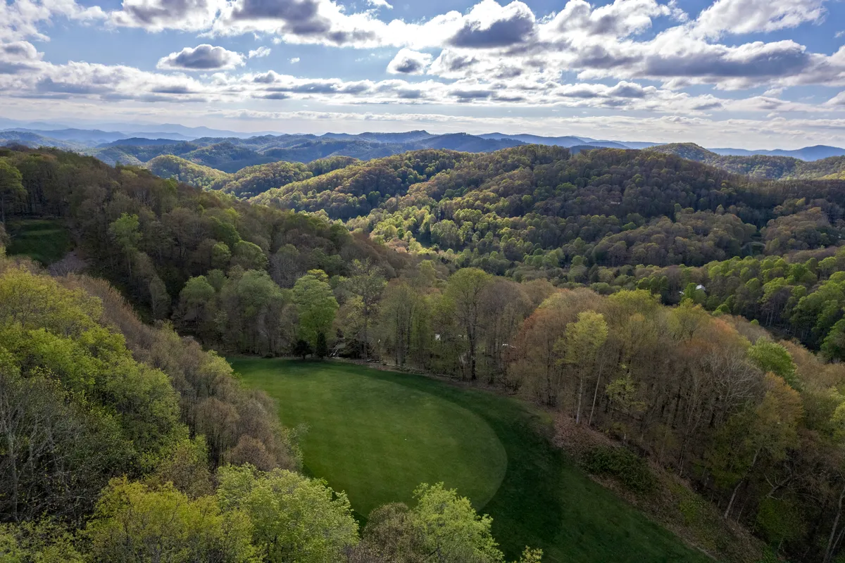 A panoramic view stretches over a lush green field with rolling hills and distant Blue Ridge mountains under a cloudy sky. The wide-open natural landscape beckons for outdoor activities like hiking, nature observation, and photography.