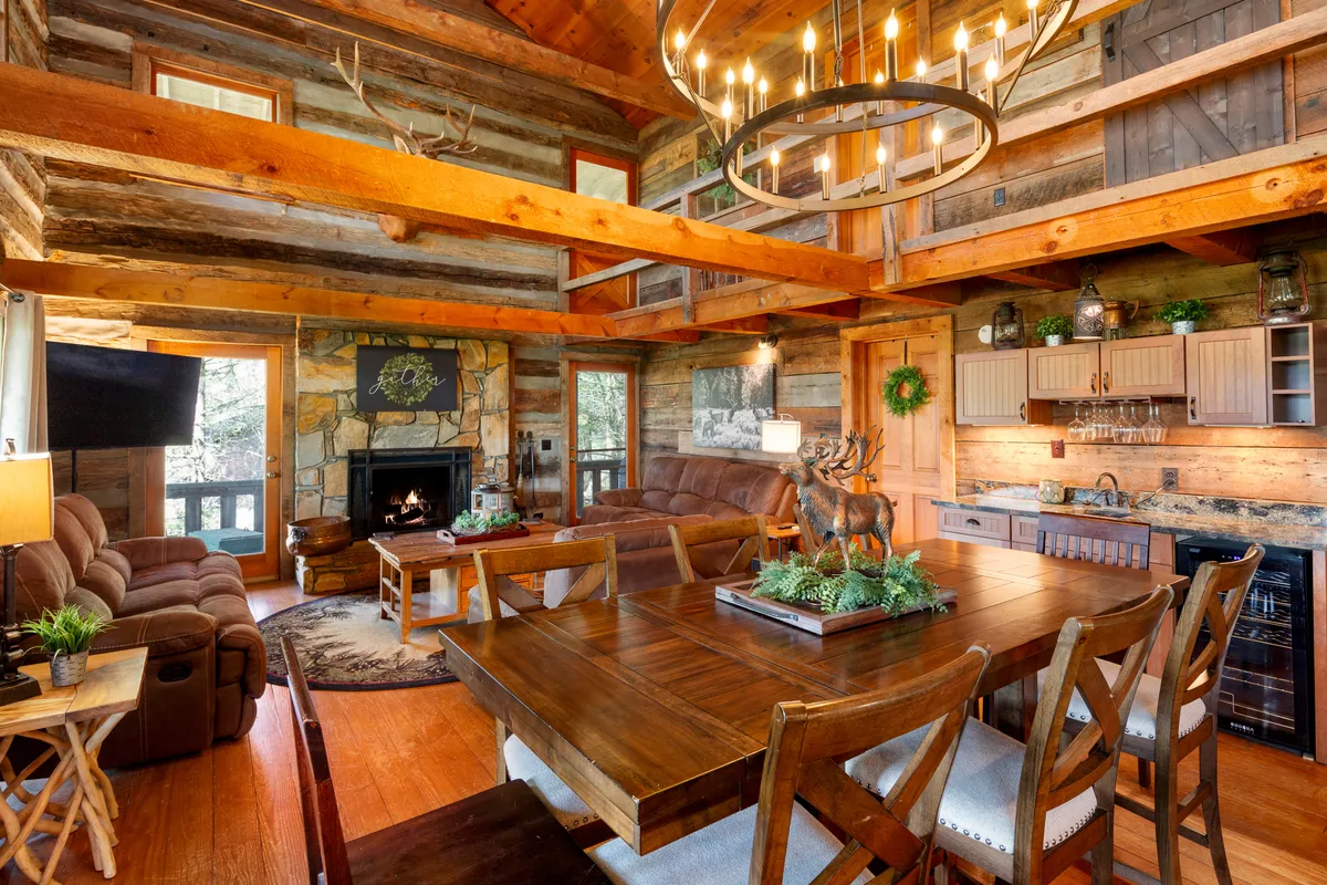 A spacious, open-plan living and dining room featuring soaring wooden beams, a stunning chandelier, a stone fireplace with a cozy fire, a plush sofa, and a grand dining table set for gatherings. The decor blends antique finishes and rustic elegance, with a decorative reindeer statue adding holiday charm.