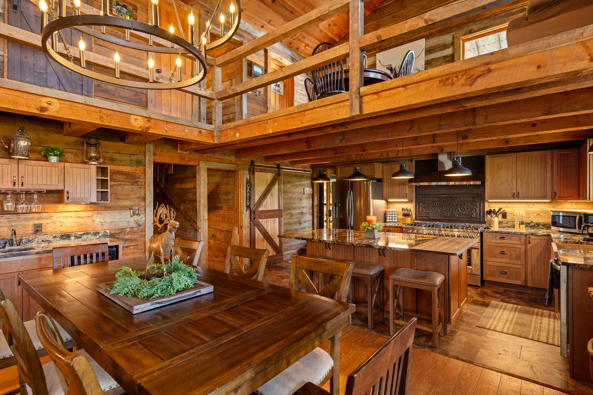 An expansive dining area with a large wooden table set for a meal, a classic chandelier overhead, exposed wooden beams, and an open kitchen layout in the background. The scene is filled with natural light, emphasizing the welcoming and communal spirit of the space.