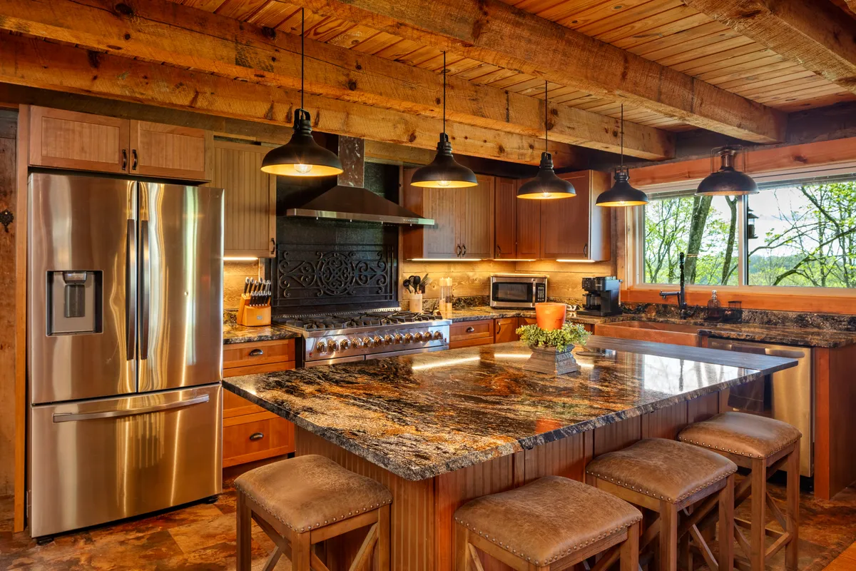 A warmly lit kitchen featuring a granite countertop with stainless steel appliances, exposed wooden beams above, pendant lights adding ambiance, and a large window that fills the space with natural light. The layout includes ample workspace for meal prep and areas for gathering, like the breakfast nook and bar seating.