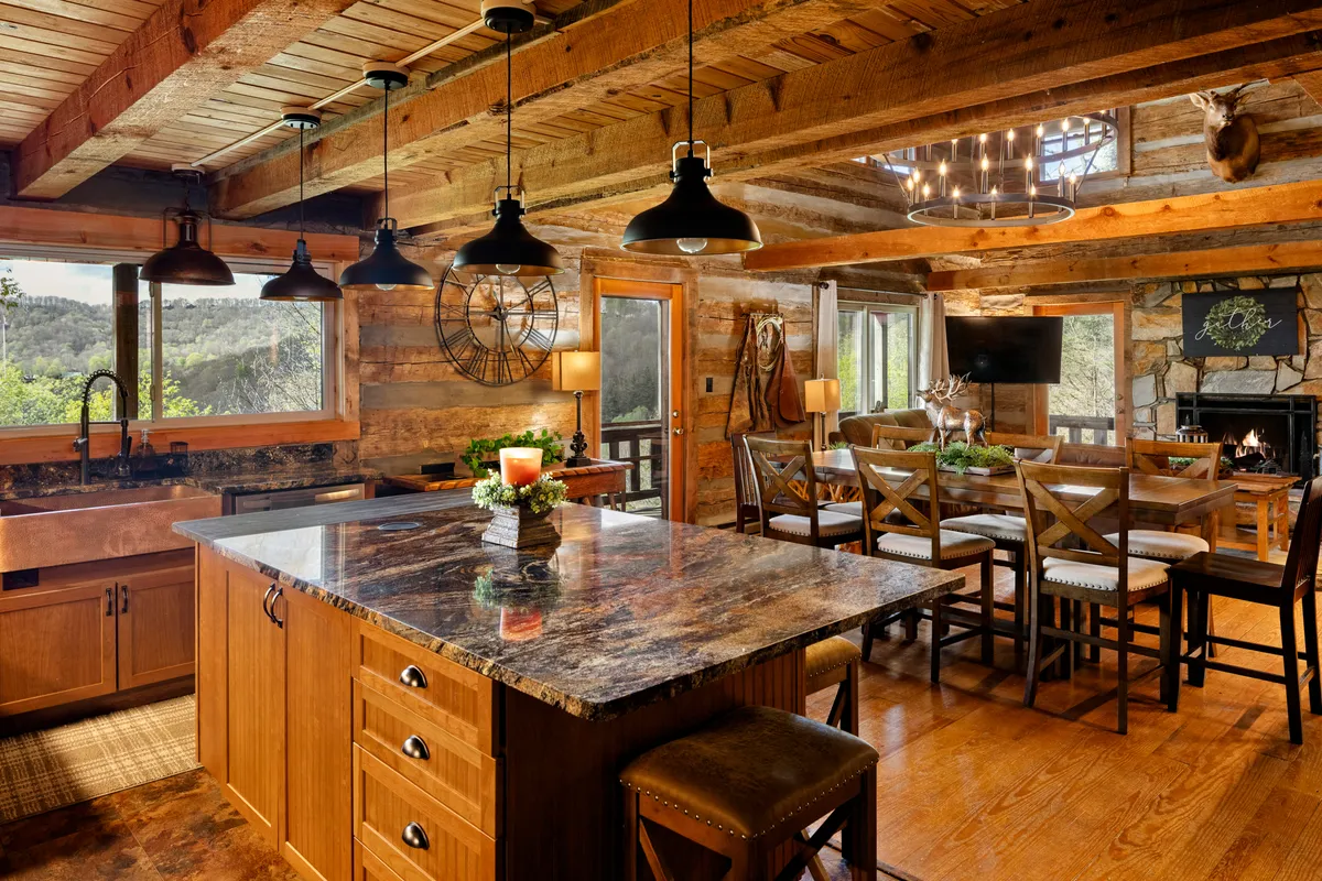 A spacious kitchen with a large island, adjoining dining table, exposed wooden beams overhead, a fireplace in view, and windows providing a beautiful natural vista. The space is set up for communal cooking, entertaining, and enjoying meals together, with a rustic yet modern aesthetic.