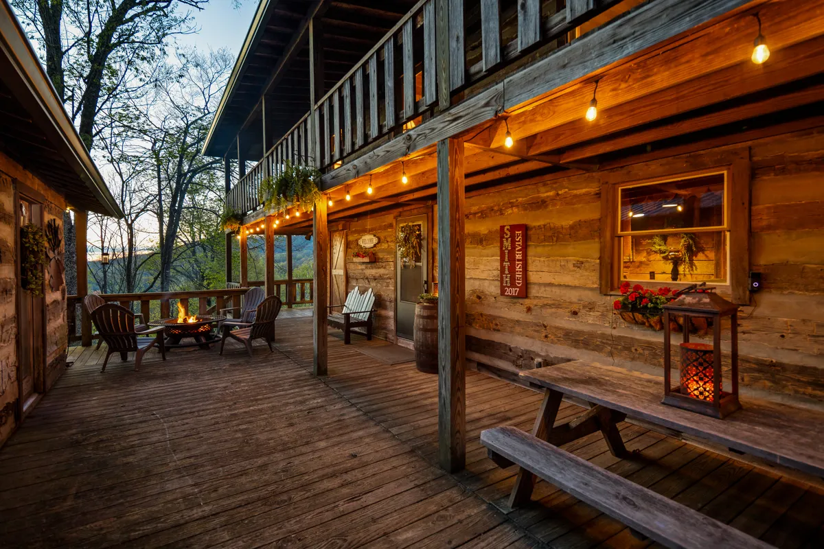 An outdoor fire pit area with wooden seating, hanging lights, and a wooden railing overlooking the outdoors. The setup suggests cozy gatherings, stargazing, and relaxing evenings in a tranquil setting.