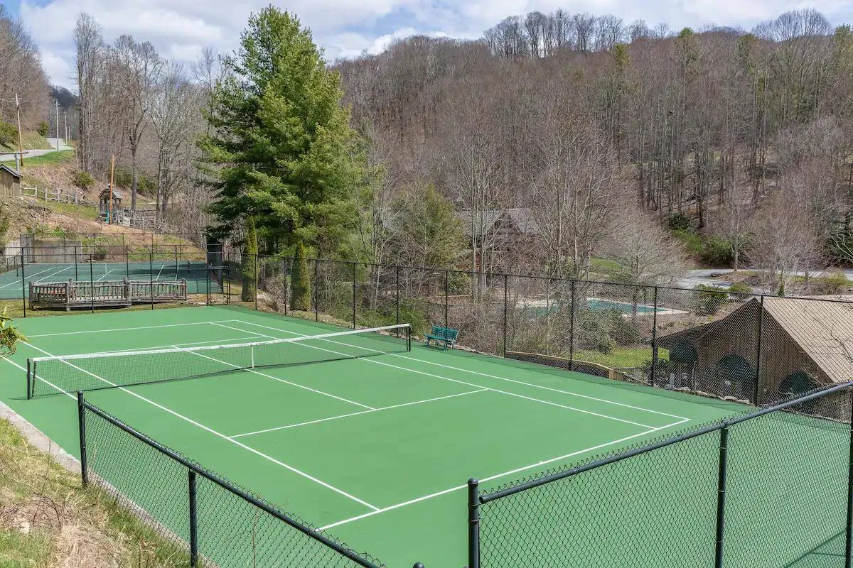 An expansive view of a tennis court nestled amid trees, with benches for relaxation and a distant glimpse of a pool area. The scene is lively and energetic, visually connecting the estate to recreational amenities and the peaceful mountain landscape.