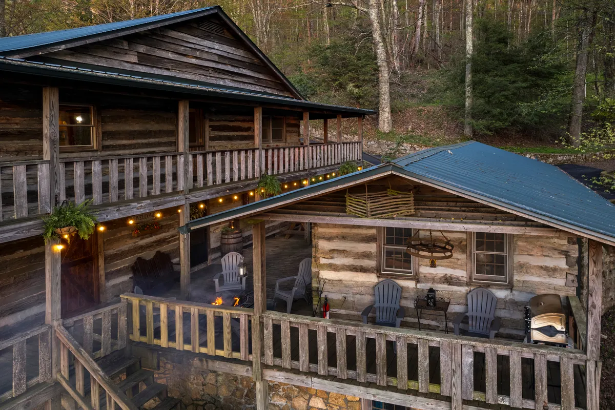 A spacious wooden cabin exterior is illuminated by warm string lights. Comfortable outdoor seating is arranged around a glowing fire pit. The wraparound deck features several spots for groups to gather, eat meals, or relax. The surrounding trees and set-back location offer privacy and immersive mountain atmosphere.