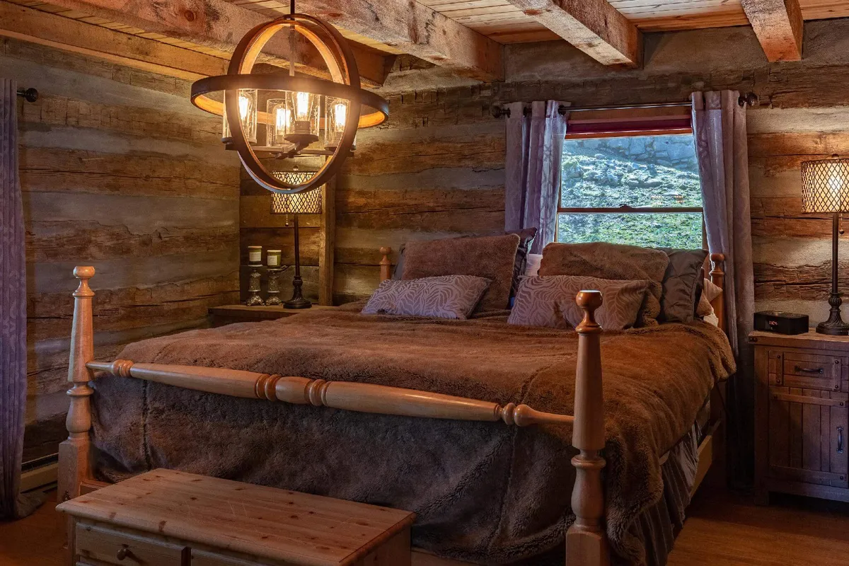 A warmly lit bedroom featuring a plush bed with luxurious bedding, a chandelier hanging from exposed wooden beams, and a window offering views of the surrounding woodland. The nightstand and decorative accents contribute to an atmosphere of comfort and tranquility.