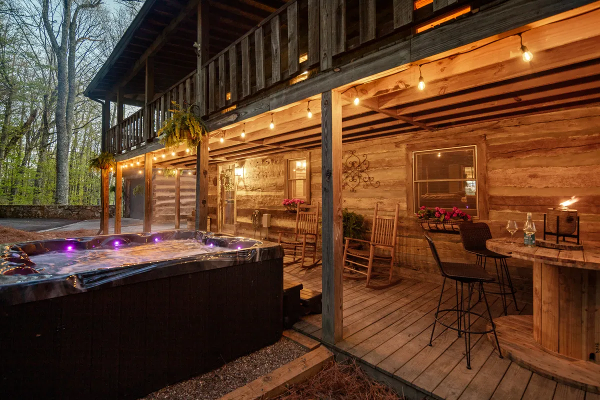 A hot tub sits on a wooden outdoor deck, with string lights overhead casting a gentle glow. Nearby, cozy seating offers a space for dining or socializing while soaking up the view. The mountains and trees in the background reinforce the connection to nature and sense of seclusion.