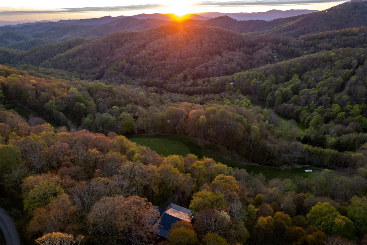 A wide-angle view capturing a dramatic sunset over a mountain range and dense forest, with the house nestled in the scene. The setting radiates natural beauty and peaceful seclusion, underscoring the property’s two-acre estate and mountain locale.