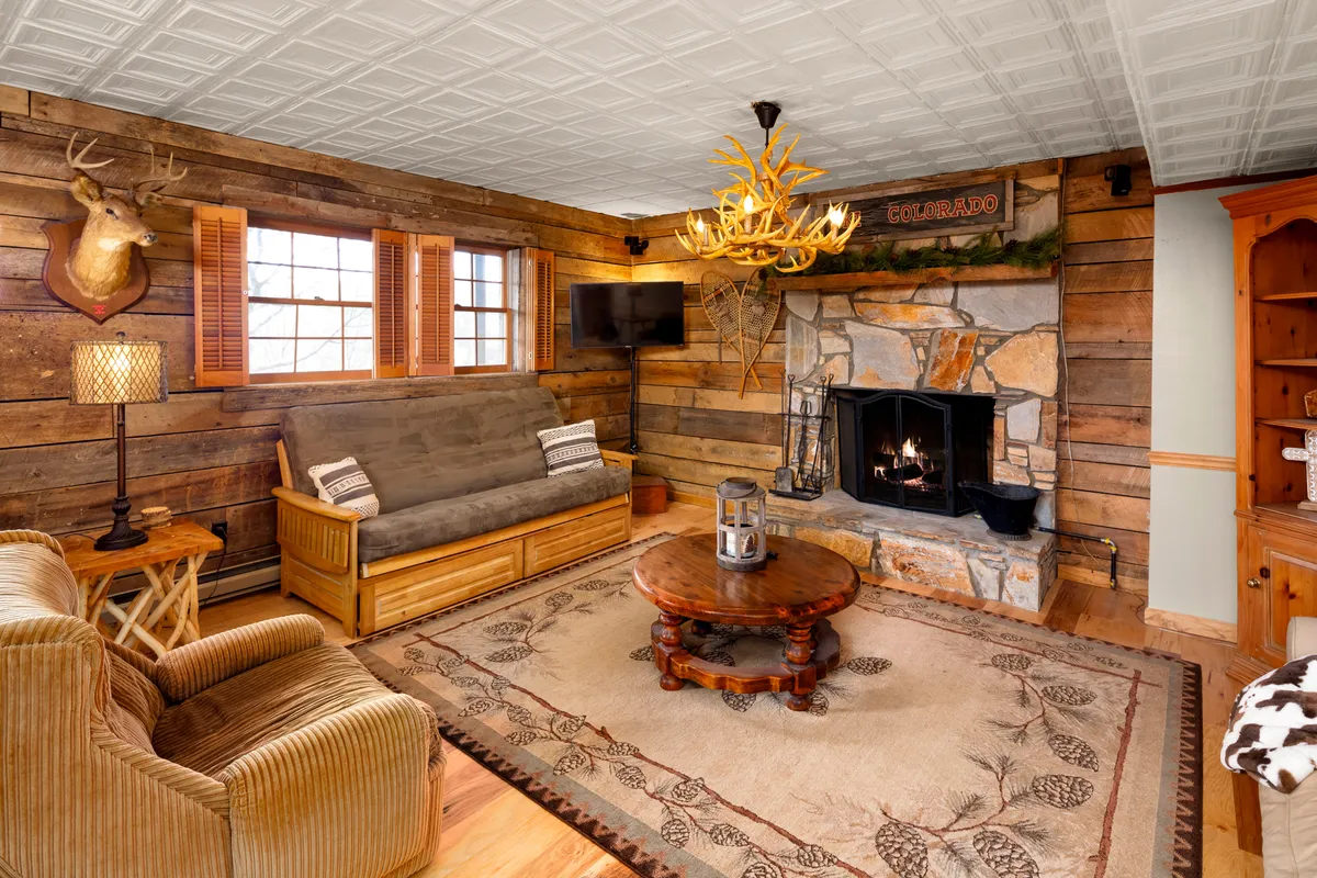 A cozy living room centered around a large stone fireplace, with an antler chandelier casting a soft glow over rustic wooden furniture and a woven rug. The space is warmly lit and inviting, perfect for family gatherings or quiet evenings.